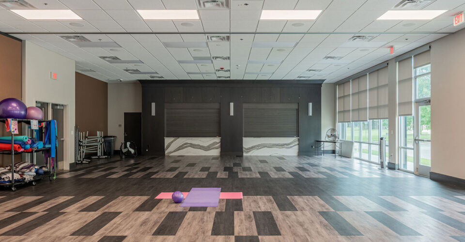 Spacious exercise room with yoga mats and an exercise ball on the floor, fitness equipment on shelves, large windows, and patterned flooring.