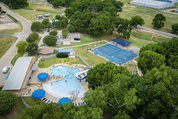 Aerial view of a recreational facility with a swimming pool, shaded lounge chairs, a basketball court, nearby buildings, trees, and parking lot.