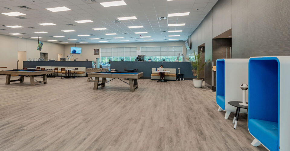 Large modern office common area with wood flooring, multiple tables, lounge pods, and a reception desk in the background under bright ceiling lights.