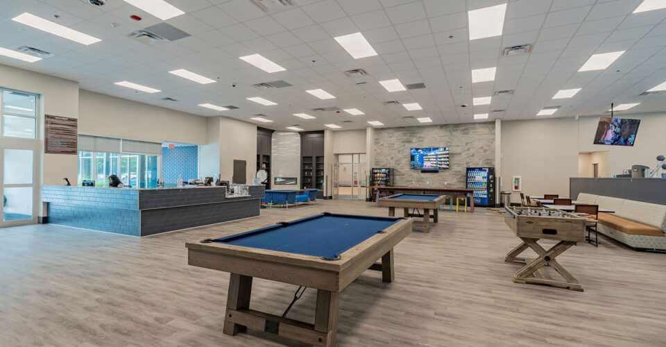A spacious rec room with pool tables, foosball, lounge seating, a TV, and a cafe area under bright ceiling lights.