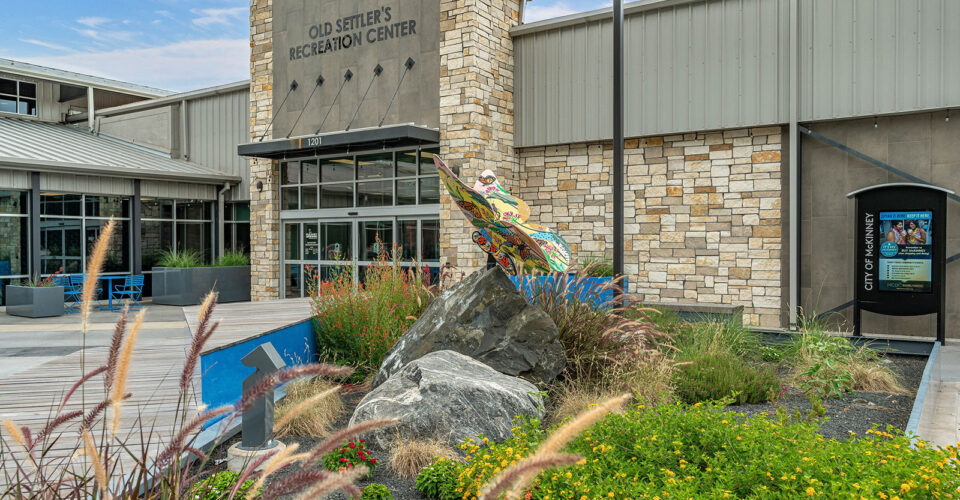 Front entrance of Old Settler's Recreation Center with a colorful fish sculpture on a rock, surrounding landscaping, and a stone and metal building exterior.