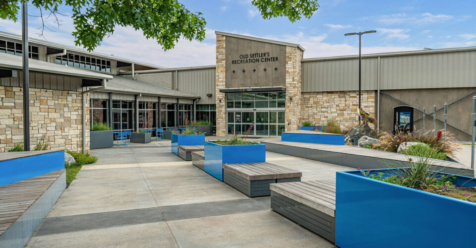 The entrance to Old Settlers Recreation Center features modern benches, blue planters, stone walls, large windows, and surrounding greenery under a blue sky.