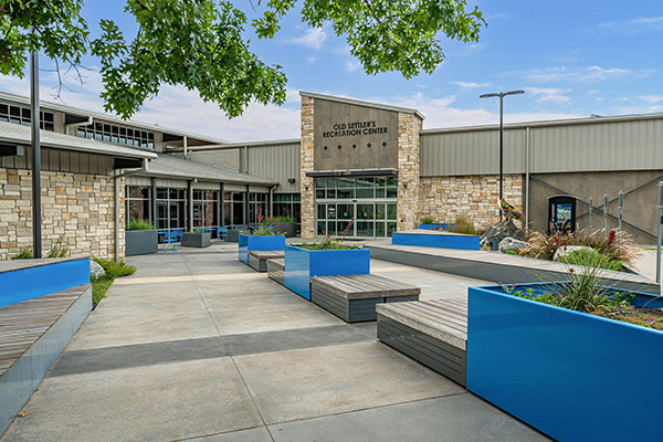 Entrance to Old Settler’s Recreation Center with stone facade, large windows, blue benches, planters, and a clear sky overhead.