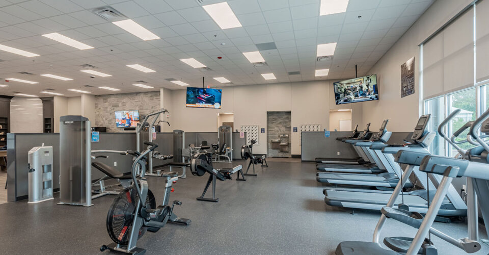 Modern gym interior with cardio machines, stationary bikes, strength equipment, flat-screen TVs, large windows, and bright overhead lighting.