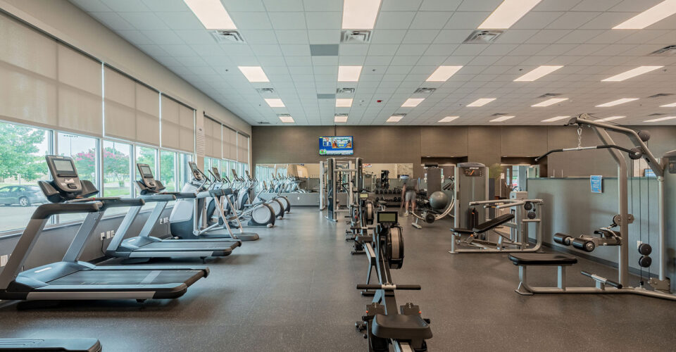 Spacious gym interior with treadmills, exercise bikes, rowing machine, weight machines, and large windows allowing natural light.