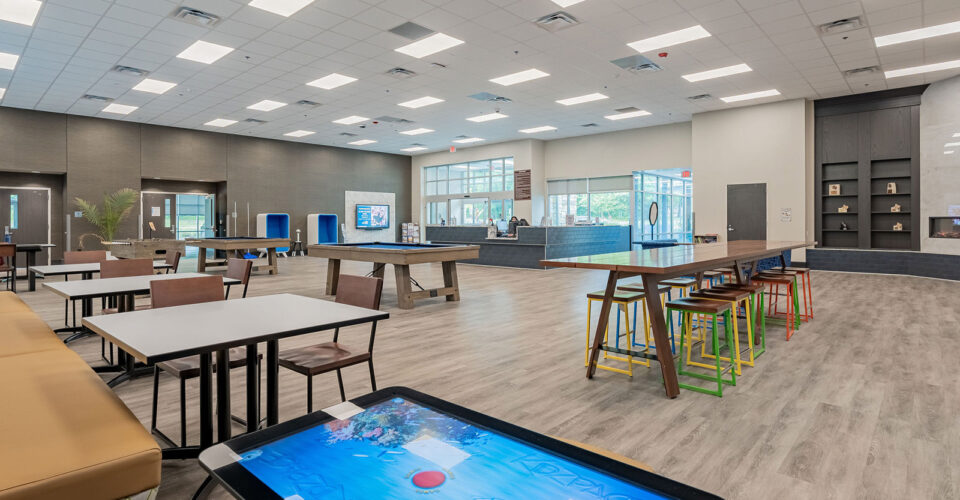 A spacious, modern common area with tables, colorful stools, a digital touchscreen, and large windows letting in natural light.