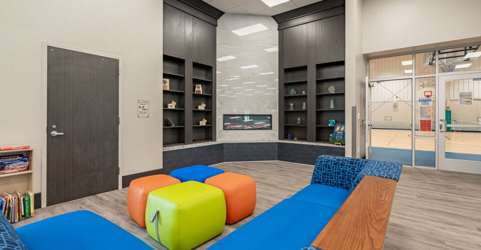 A modern lounge area with colorful cube seats, blue cushioned benches, built-in shelves, and a view into a gymnasium through double glass doors.