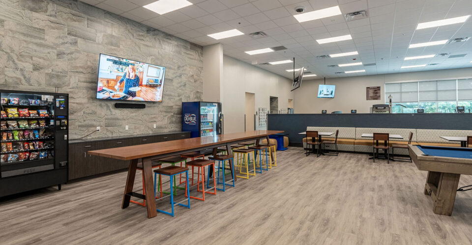 Modern break room with snack and drink vending machines, a long wooden table with colorful stools, booth seating, and a pool table; televisions are mounted on the walls.