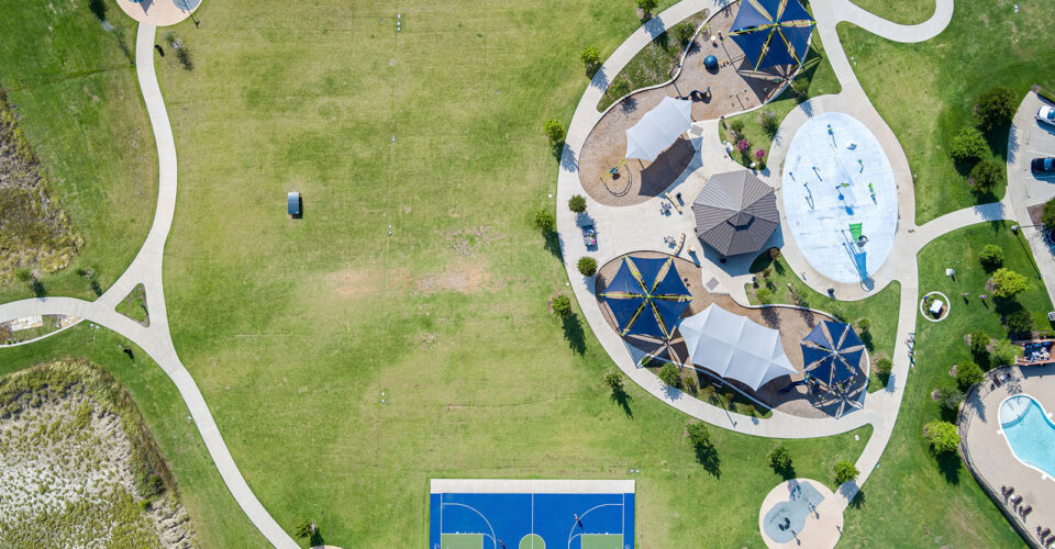 Aerial view of a park with walking paths, shaded picnic areas, a playground, a splash pad, a basketball court, and a swimming pool.