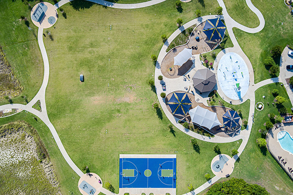 Aerial view of a park with walking paths, playgrounds, shaded picnic areas, a blue basketball court, and a nearby swimming pool.
