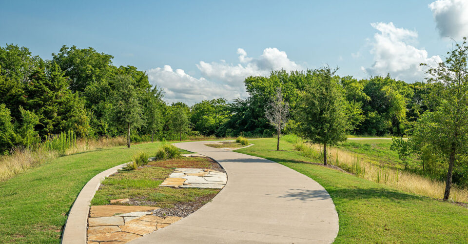 Curved concrete walking path bordered by grass, trees, and a stone-lined section under a clear blue sky with scattered clouds.