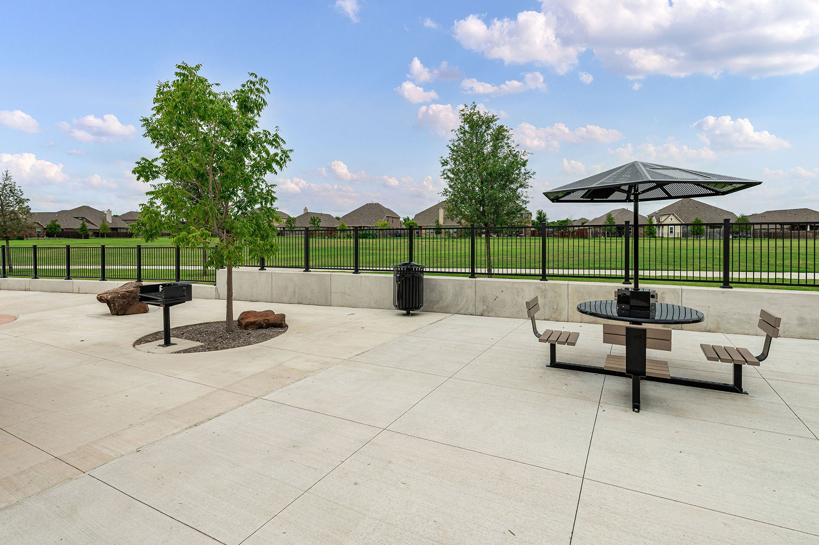 A patio area with a picnic table, umbrella, grill, trees, trash can, and a black metal fence overlooking a grassy field and houses in the background.