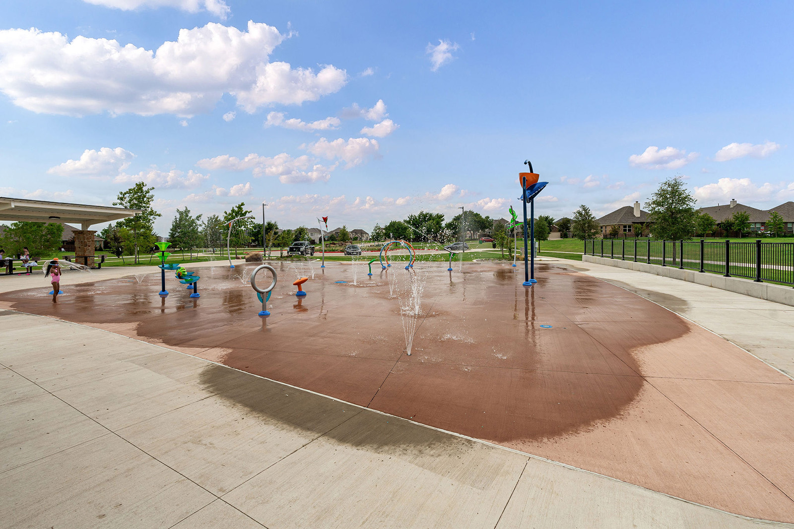 A splash pad with various water features and sprinklers sits in an open park area with grass, trees, and nearby houses under a partly cloudy sky.