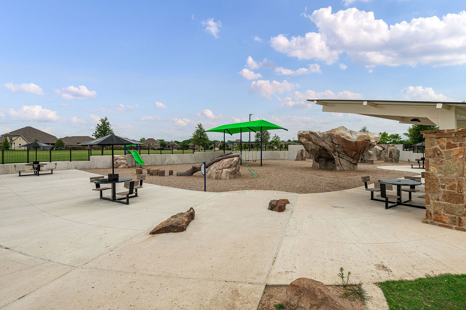 A playground area with climbing rocks, a green slide, shaded seating, picnic tables, and open space under a partly cloudy sky.