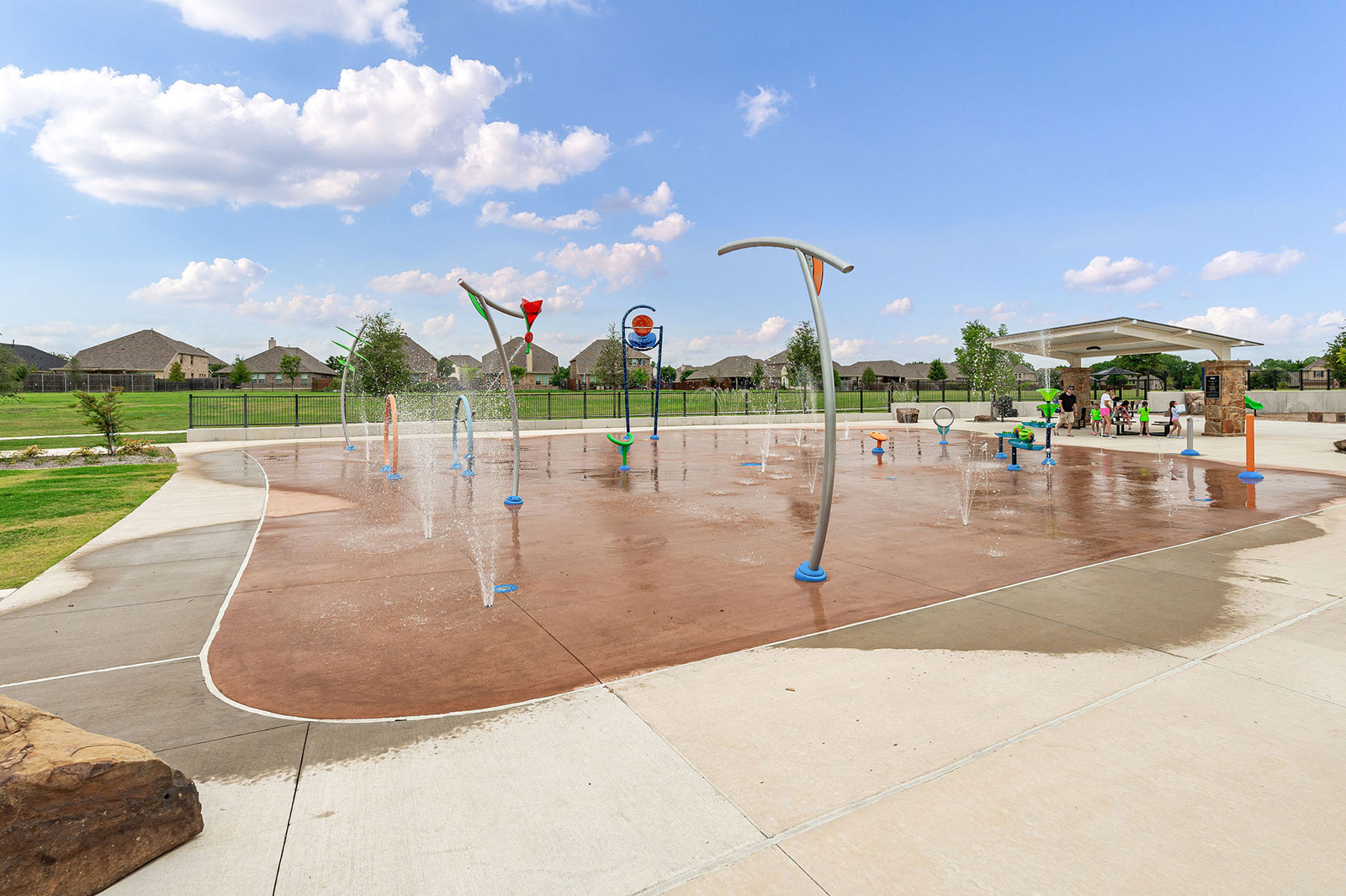 A splash pad with various water features in a park on a sunny day, with houses and a covered seating area in the background.