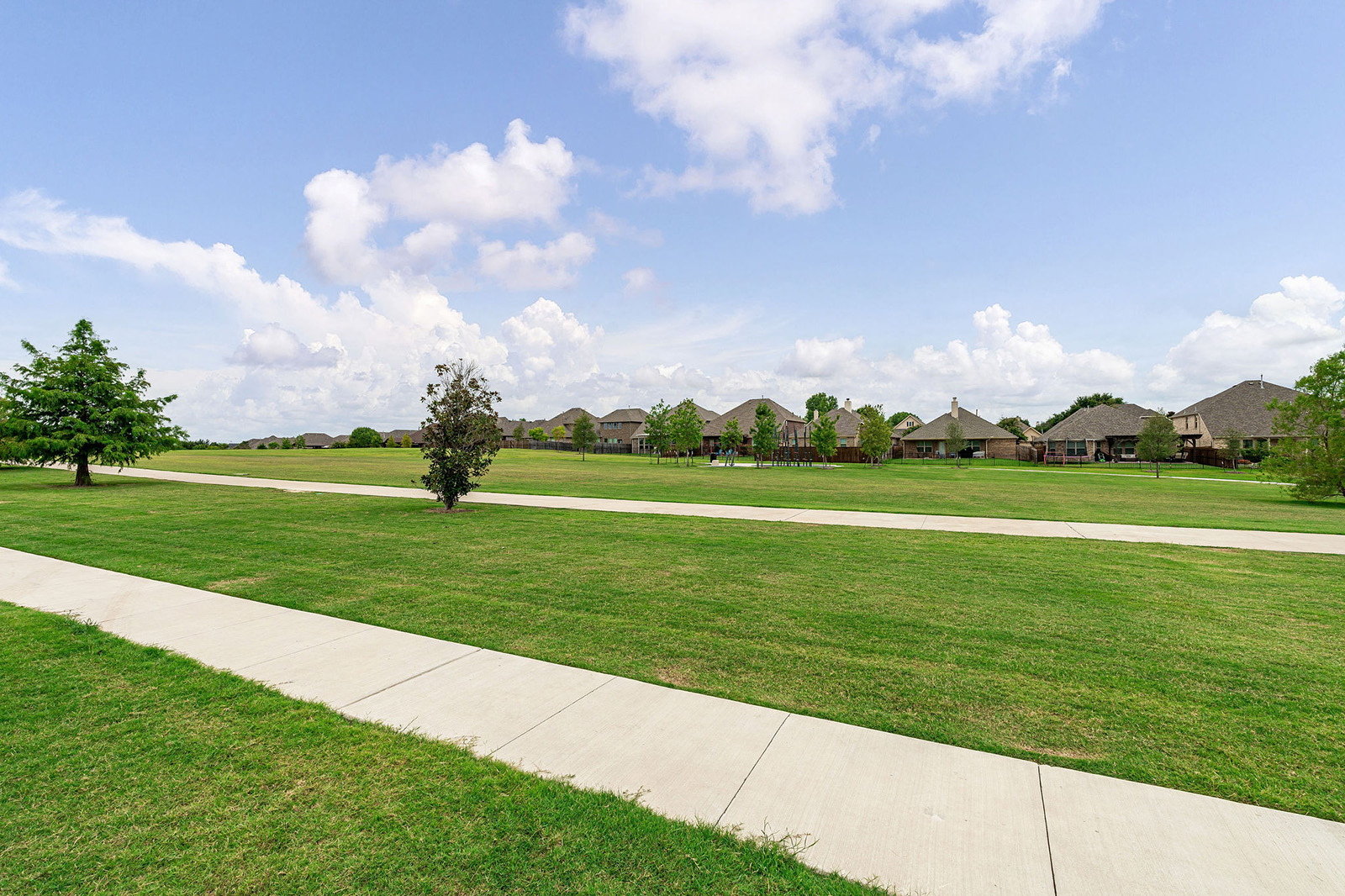 Sidewalk running alongside a wide grassy field with scattered trees, under a partly cloudy sky; houses are visible in the distance.