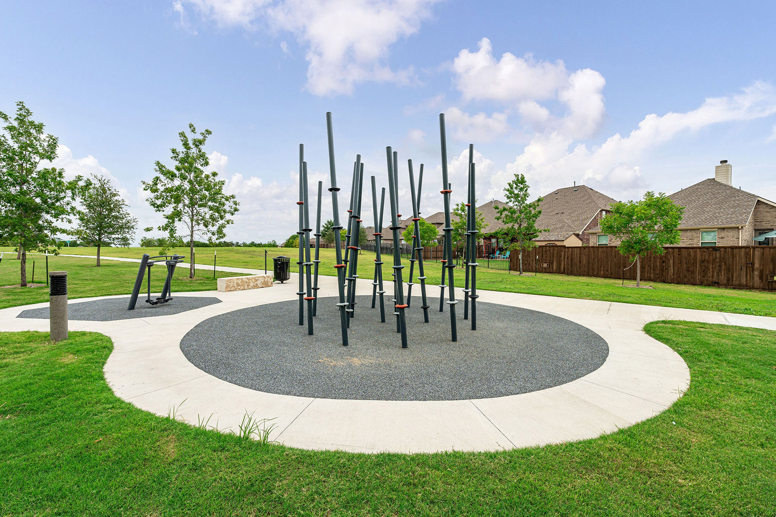 Outdoor park with metal climbing poles on a circular rubber surface, exercise equipment to the left, and houses with lawns in the background under a partly cloudy sky.