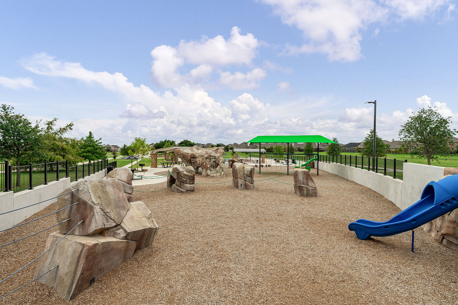 Outdoor playground with artificial rock climbing structures, a blue slide, green shade canopy, and a gravel surface, surrounded by a white wall and fence.