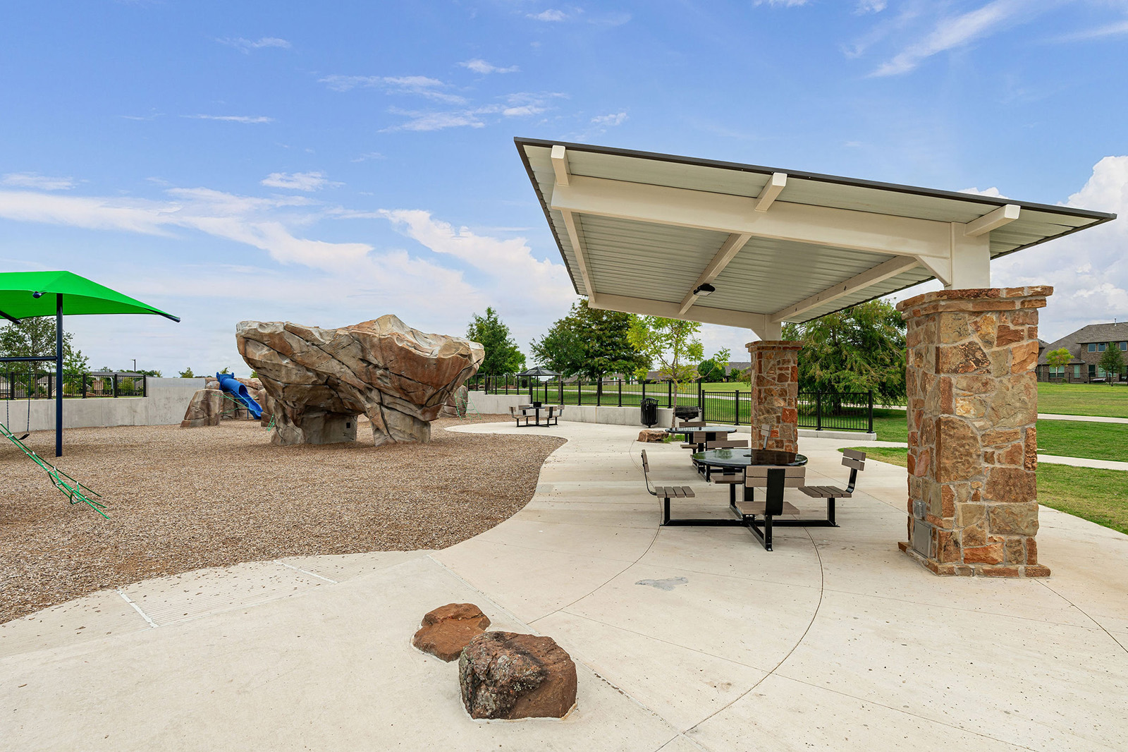 A covered picnic area with stone pillars stands near a playground with a climbing rock and slide in an open park setting.