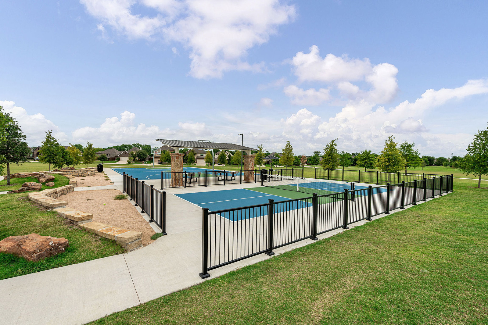 Outdoor fenced pickleball courts with surrounding green grass, picnic tables under a shelter, and a sidewalk leading into a park area under a partly cloudy sky.