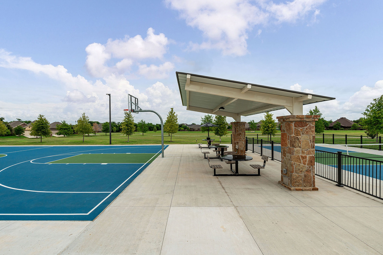 Outdoor basketball court next to a covered picnic area with tables and stone pillars, surrounded by grass and trees under a partly cloudy sky.