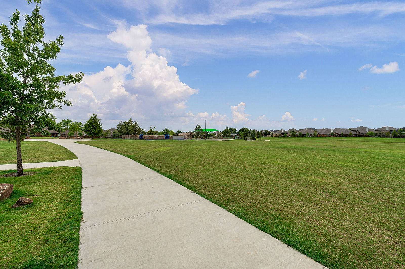 A concrete sidewalk curves through a grassy park area with scattered trees, under a partly cloudy blue sky. Houses are visible in the distance.
