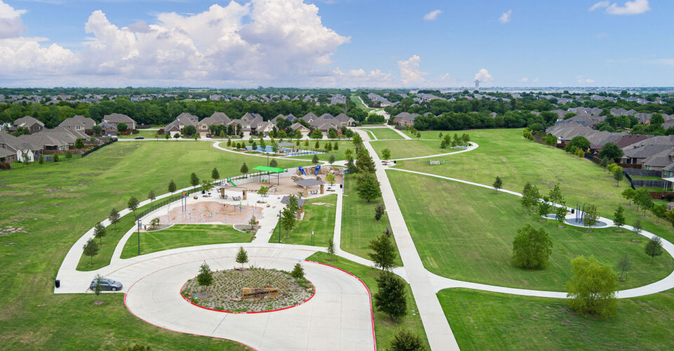 Aerial view of a suburban park with winding sidewalks, a playground, splash pad, grassy fields, trees, and surrounding houses under a partly cloudy sky.
