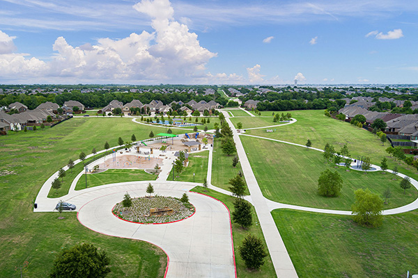 Aerial view of a suburban park with walking paths, open green spaces, a playground, and surrounding houses under a partly cloudy sky.