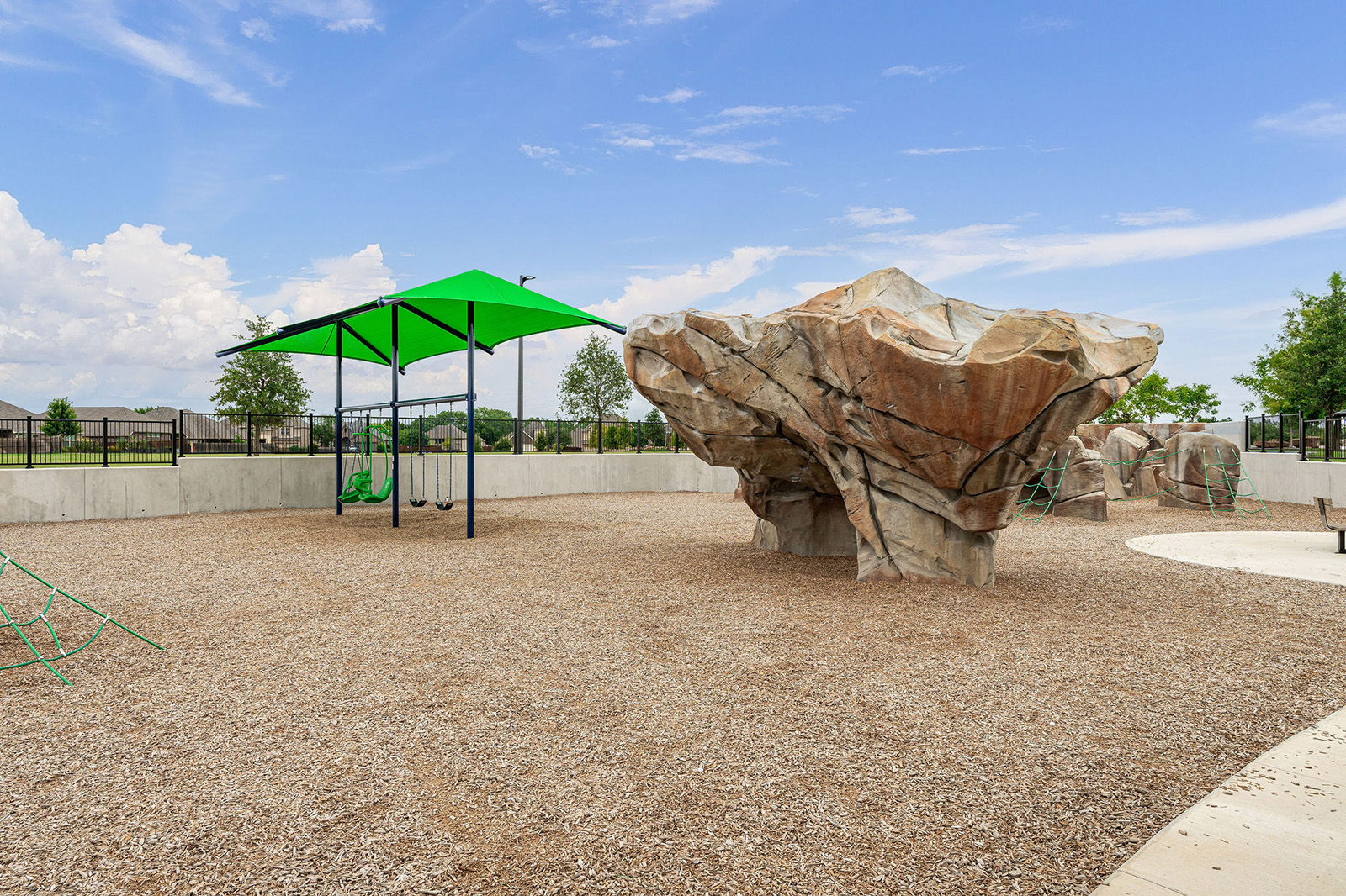 Outdoor playground with climbing rock structure, two green swings under a canopy, mulch ground covering, and a concrete border. Blue sky and suburban homes in the background.