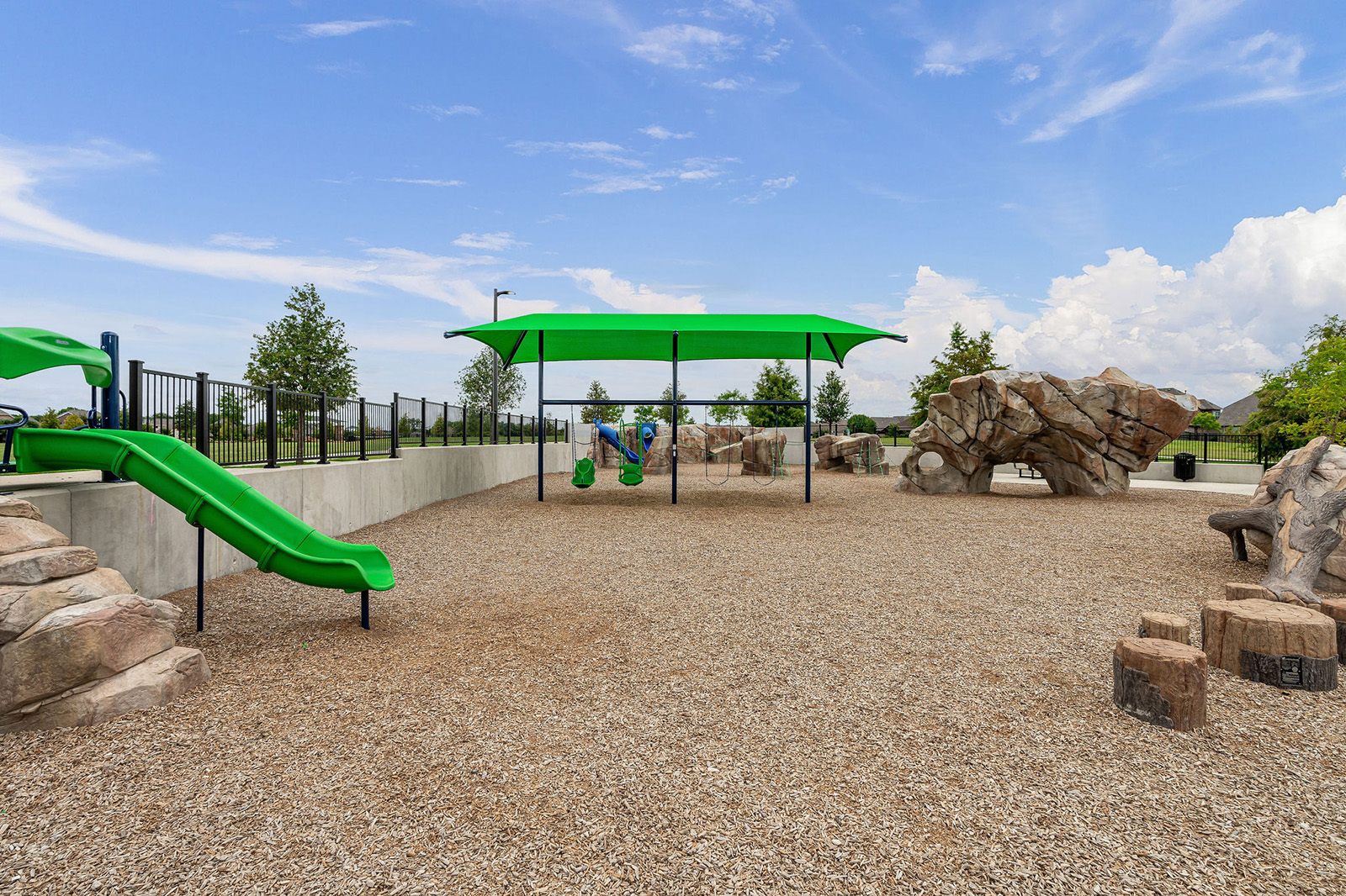 Outdoor playground with green slides, swings under a shade, artificial climbing rocks, and wood chip ground covering on a clear day.