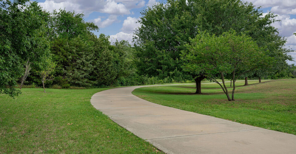 A curved concrete pathway runs through a grassy park area with scattered trees under a partly cloudy sky.