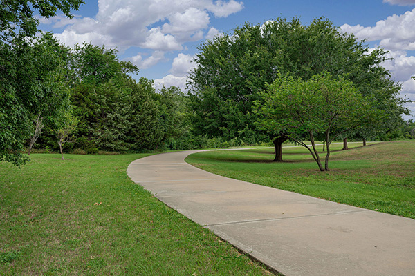 A paved walking path curves through a grassy park with scattered trees under a partly cloudy sky.