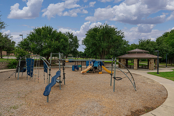 A playground with climbing structures and slides sits on gravel near a curved sidewalk, trees, and a covered picnic pavilion under a partly cloudy sky.