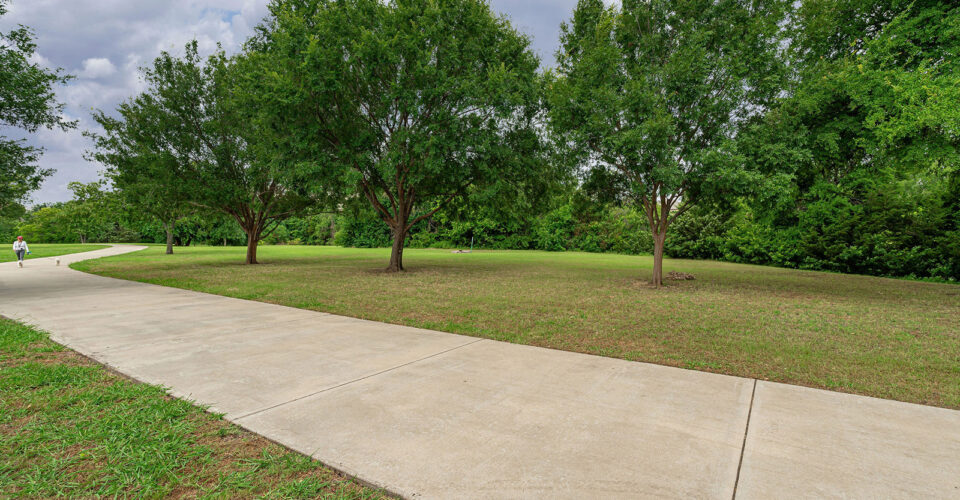 Curved concrete pathway runs through a grassy park area with several green trees and a person walking in the distance.