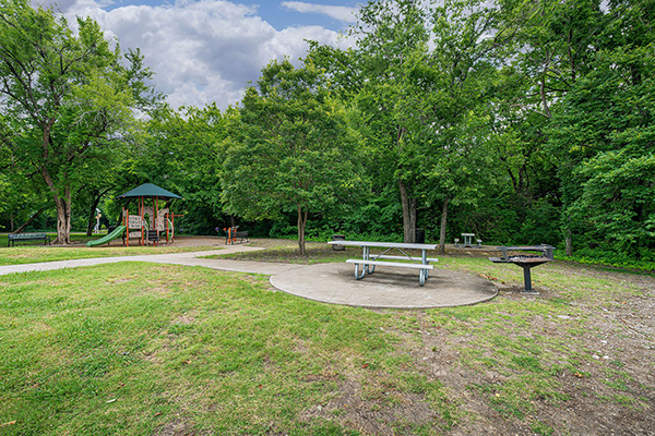 A picnic area with a metal table, a grill, and a gazebo surrounded by green trees and grass under a partly cloudy sky.