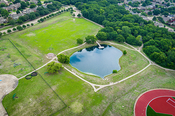 Aerial view of a park with a pond, walking paths, open grassy areas, a playground, and a red running track, surrounded by trees and residential houses.