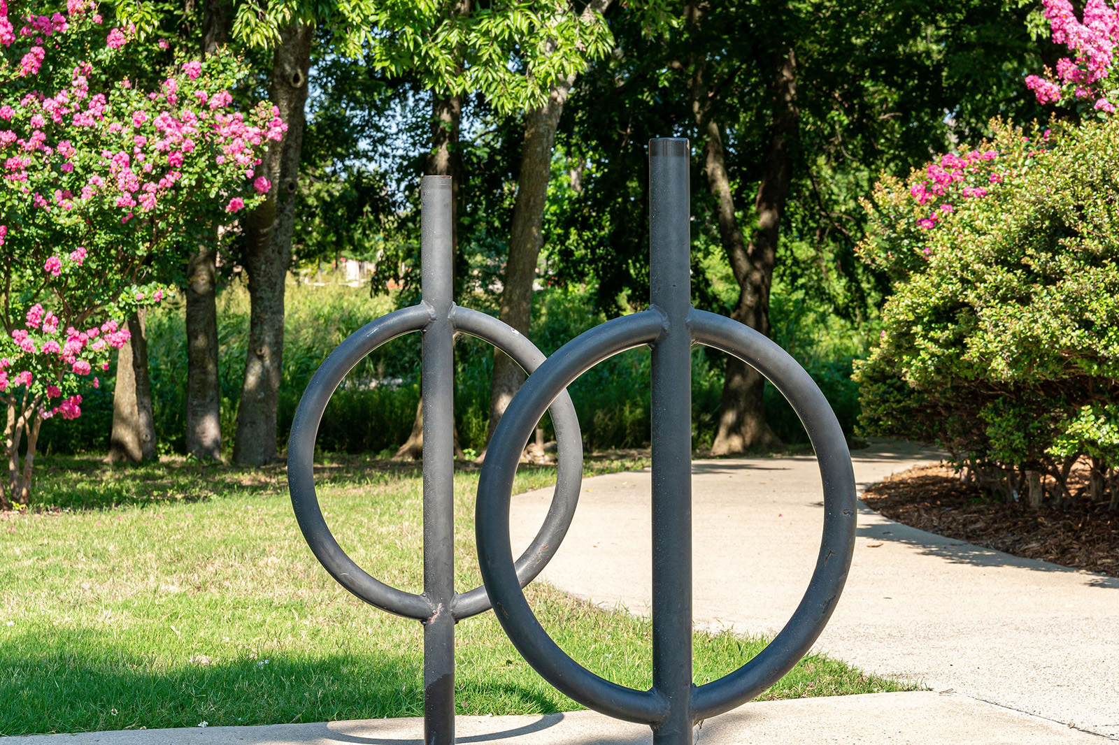 Two black circular metal bike racks are installed on a concrete pathway in a park with green grass, trees, and pink flowering bushes in the background.