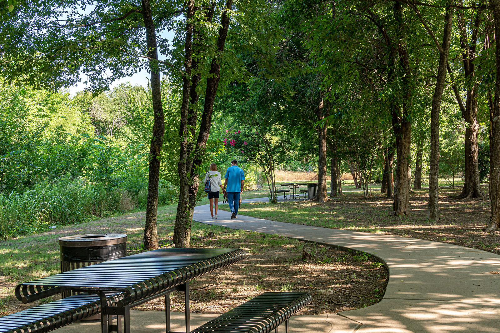 Two people walk along a winding paved path through a wooded park area with benches and picnic tables on a sunny day.