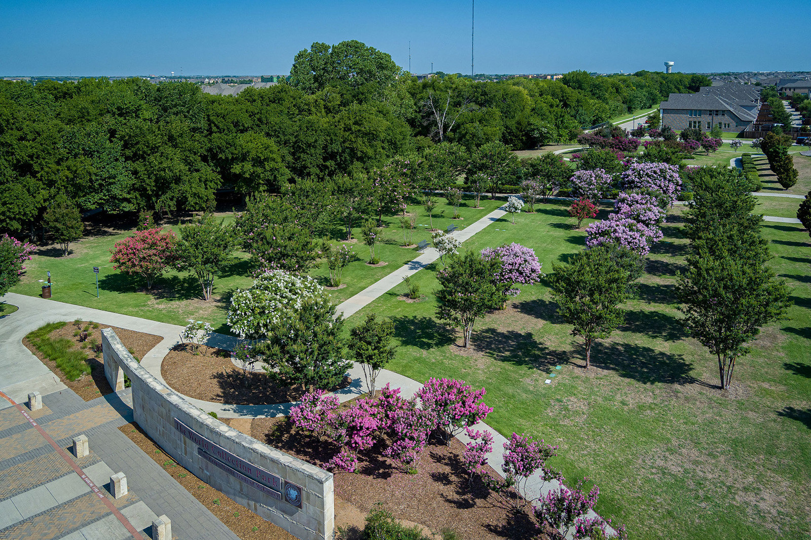 Aerial view of a landscaped park with walking paths, green lawns, and trees with pink, white, and purple flowers, surrounded by wooded areas and buildings in the background.