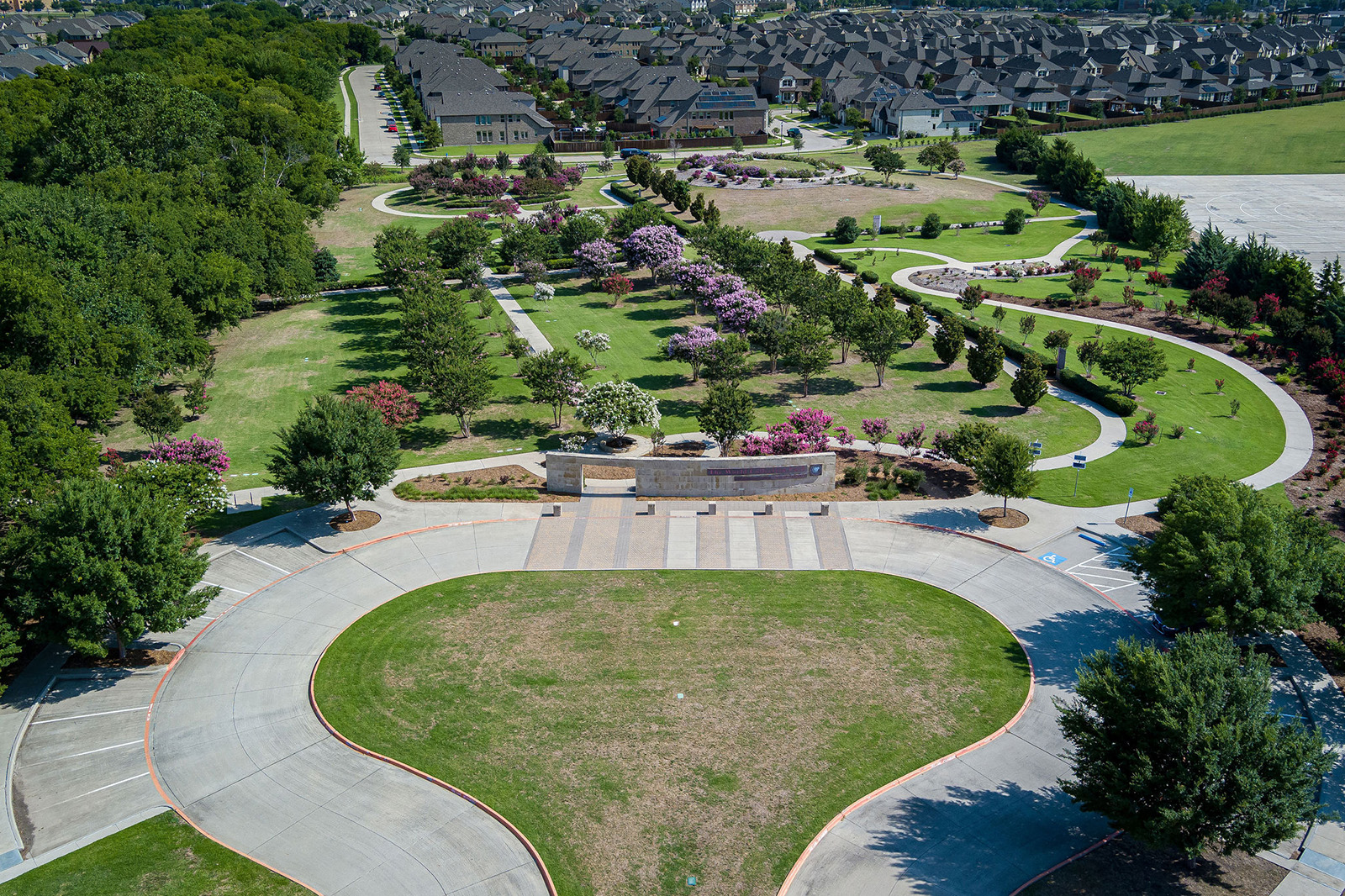 Aerial view of a landscaped park with walking paths, gardens, and a stone wall, surrounded by trees and residential houses in the background.