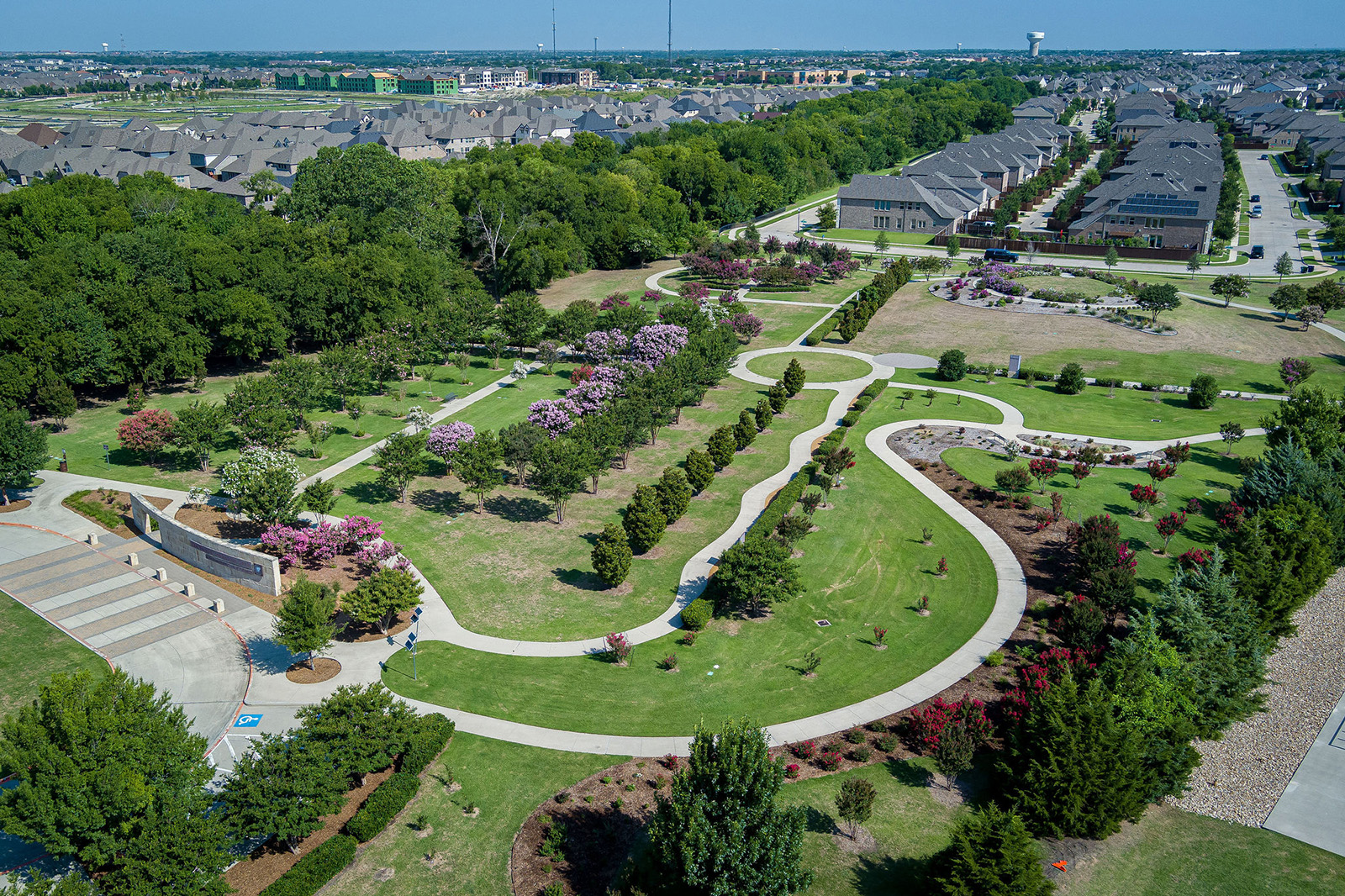 Aerial view of a landscaped park with curved walking paths, flowering trees, lawns, and surrounded by residential houses.