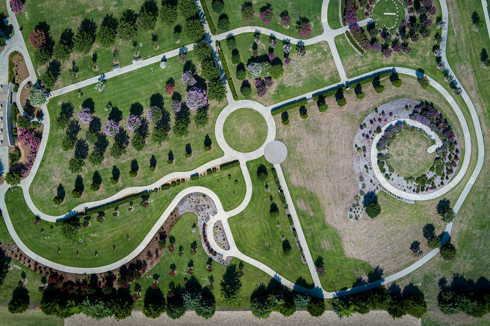Aerial view of a park with winding concrete pathways, circular plaza, landscaped trees, grassy areas, and flowerbeds.