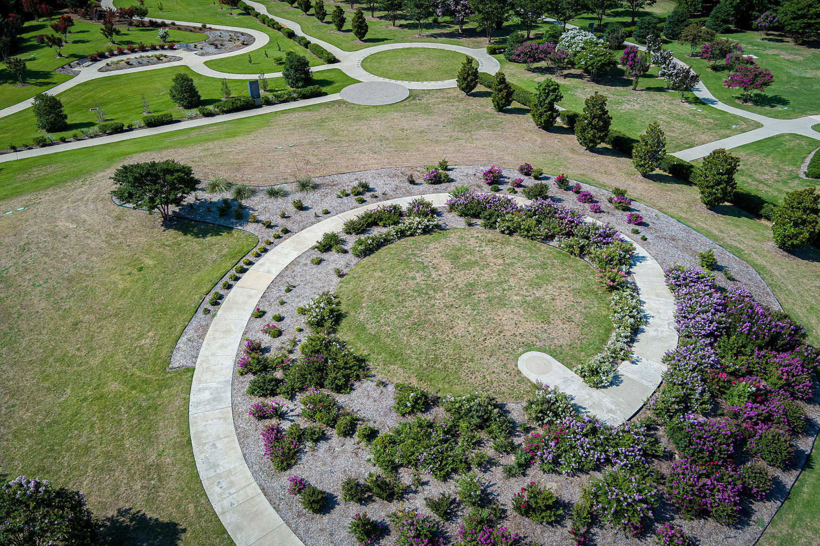 Aerial view of a landscaped park with circular pathways, green lawns, and flower beds, featuring a prominent spiral-shaped garden area.