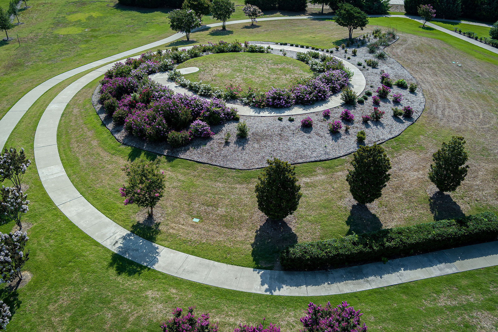 Aerial view of a circular walking path surrounding landscaped flower beds and trees in a grassy park area.