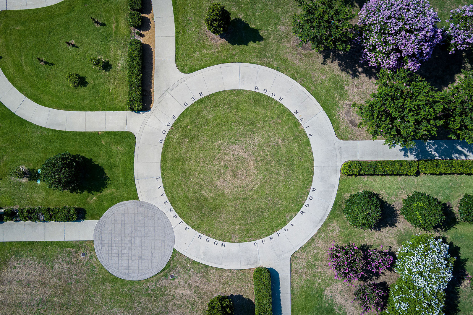 Aerial view of a circular concrete walkway with engraved text on the edge, surrounded by grass, trees, and intersecting pathways in a landscaped area.