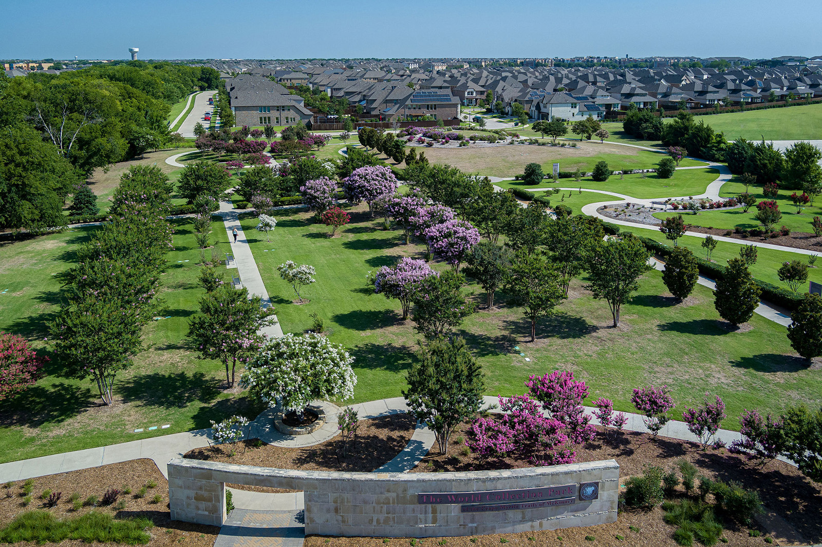 Aerial view of a landscaped park with walking paths, trees, blooming flowers, and residential houses in the background under a clear sky.