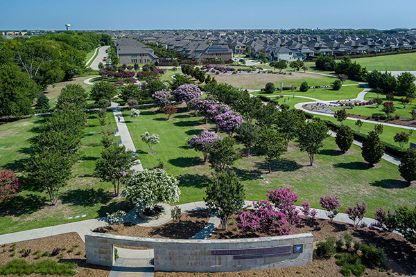Aerial view of a landscaped park with walking paths, blooming trees, and open green spaces, surrounded by residential houses in the background.