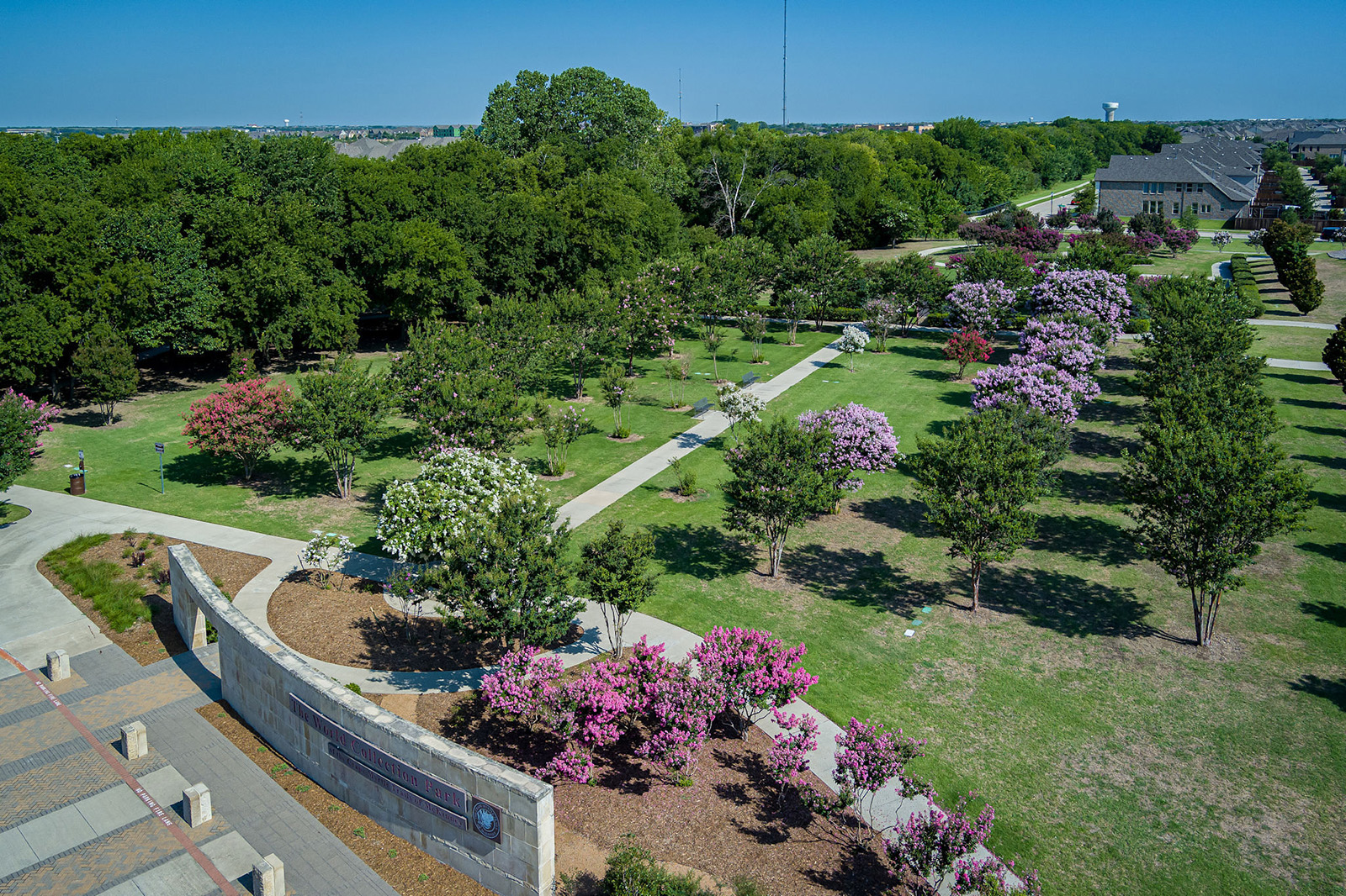 Aerial view of a park with walking paths, green grass, various trees, and blooming pink and white flowering trees under a clear sky.