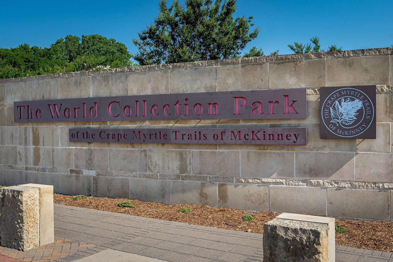 A stone wall sign reads "The World Collection Park of the Crape Myrtle Trails of McKinney" with a circular park logo on the right side.