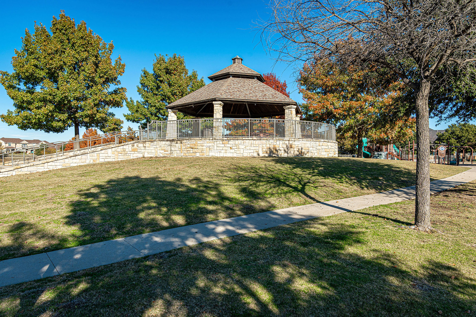 Stone gazebo with a shingled roof sits on a small hill surrounded by trees and grass, with a paved walkway leading up to it under a clear blue sky.